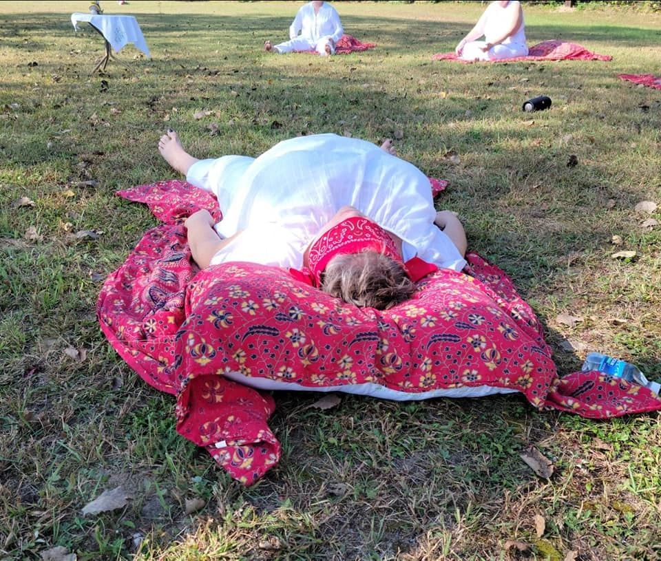 A participant dressed in white resting on a red blanket outdoors after ceremony, with others seated in the background